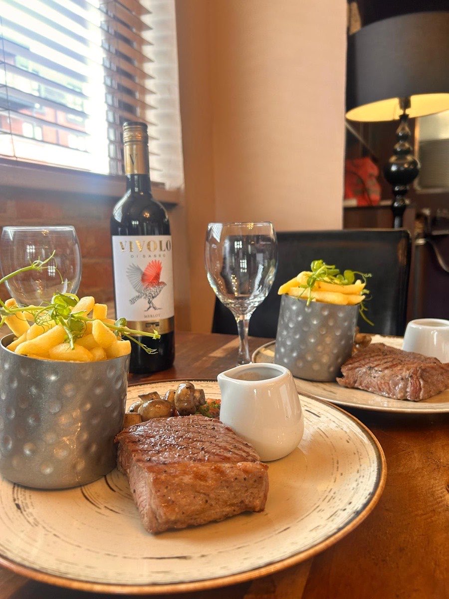 Steak with chips in a tin cup, mushrooms and a jug of sauce, with a bottle of red wine on the table